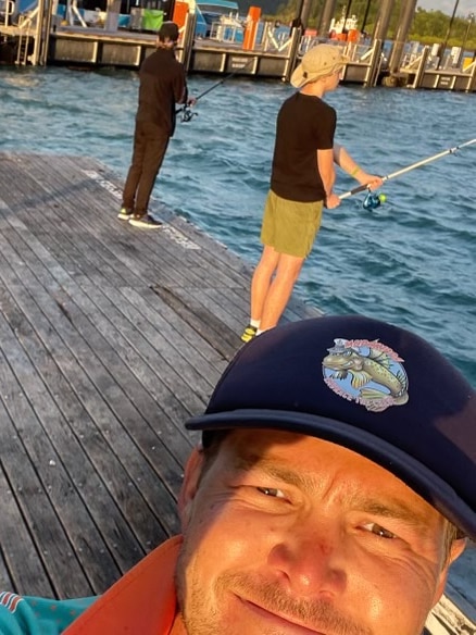 A man in the foreground takes a selfie with two boys behind him fishing off a jetty. 