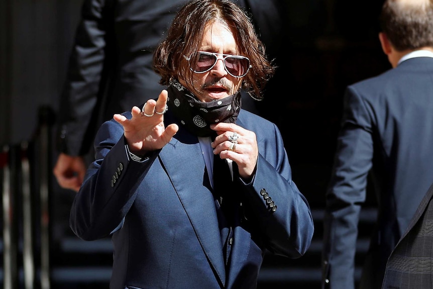Actor Johnny Depp wears white sunglasses and pulls down a black mask while wearing a navy suit.