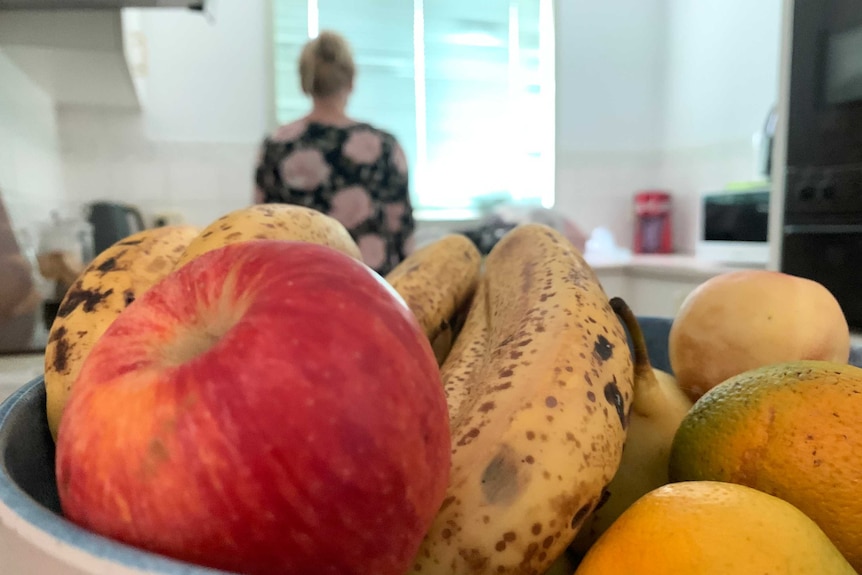 A fruit bowl in foreground, with an out of focus woman standing in a kitchen in background.