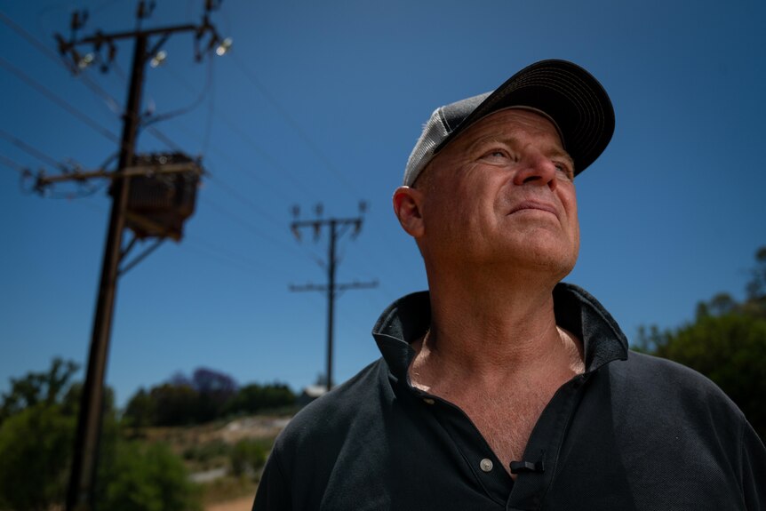 A man with a baseball cap and serious expression looks off camera, powerlines behind him