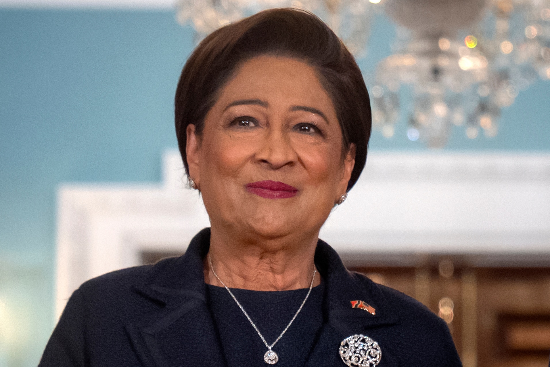 Kamla Persad-Bissessar wearing a dark jacket, silver necklace and brooch standing while smiling in a turquoise room