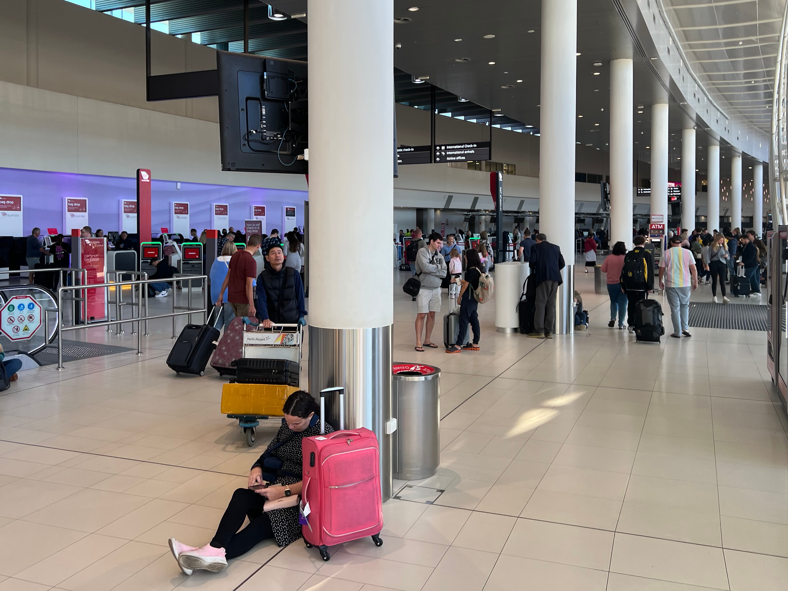 Passengers mill about an airport terminal, with one in the foreground siutting on the floor beside a pink suitcase.