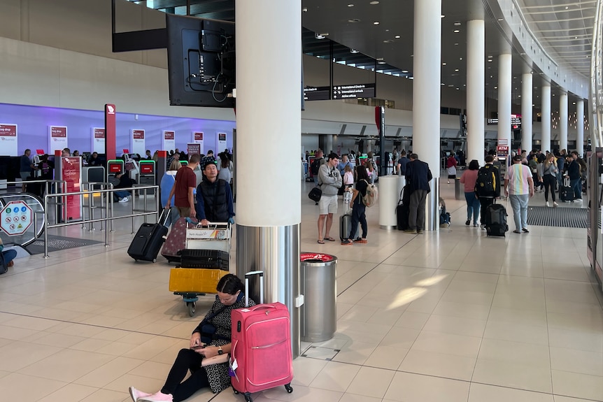 乘客们在机场航站楼周围走动，其中一位乘客坐在前景的地板上，旁边有一个粉色的手提箱。
