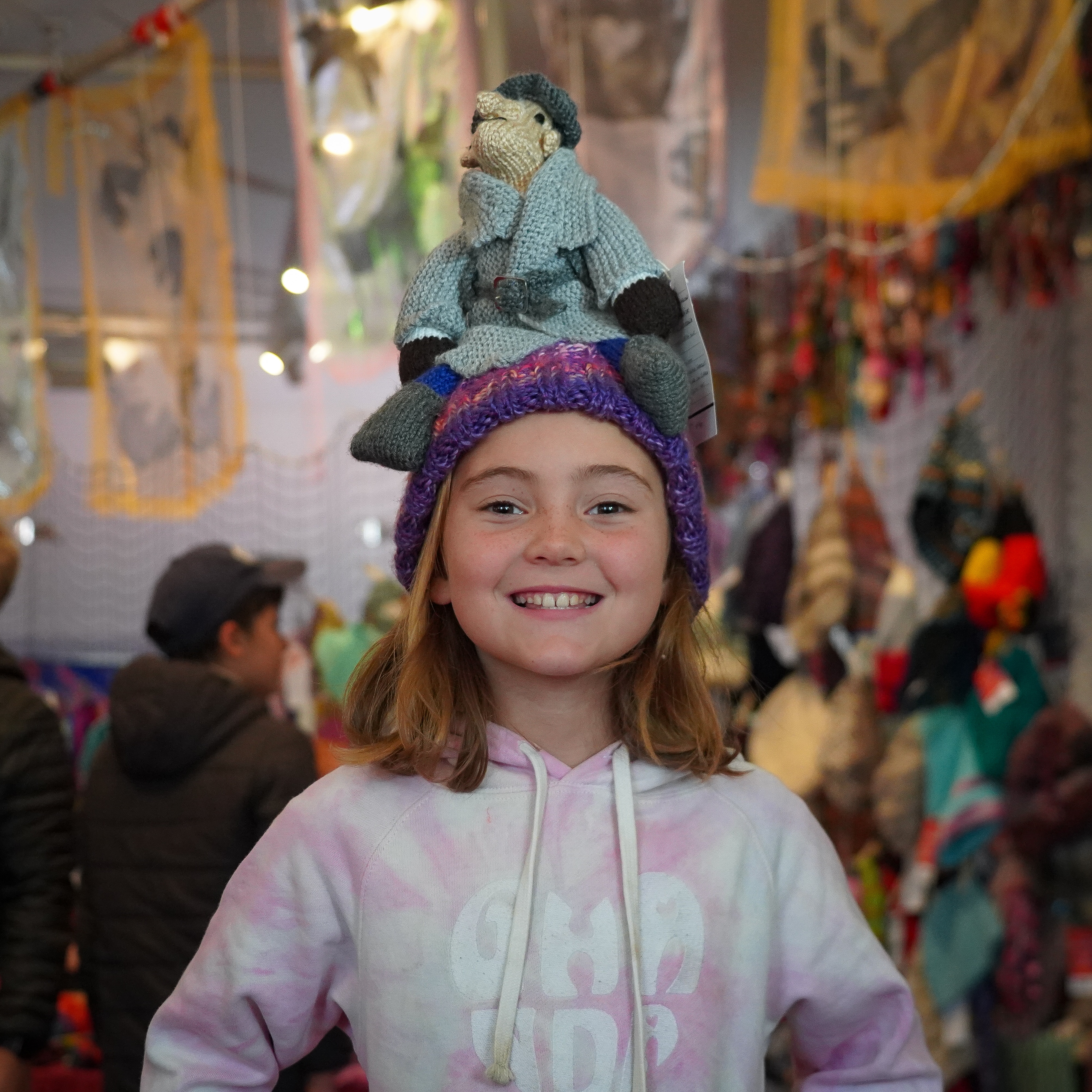 A young girl smiling at the camera, she is wearing a handmade beanie with a character perched on top of the hat.