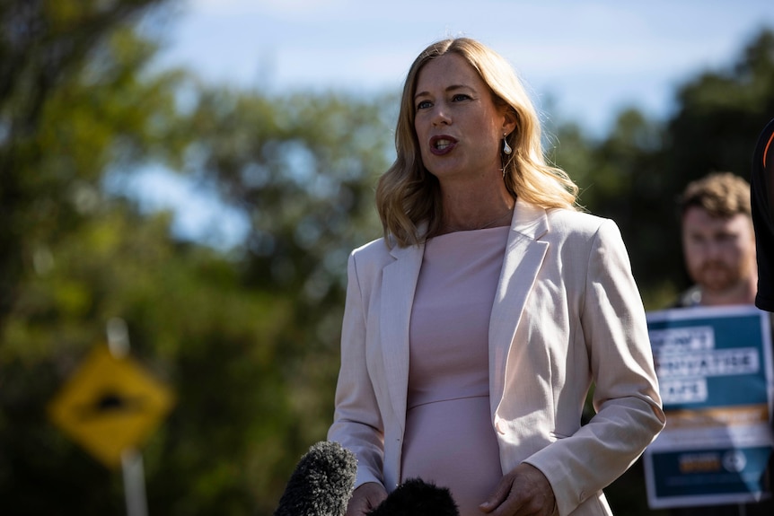 A woman speaking, wearing a white blazer and a pink shirt.