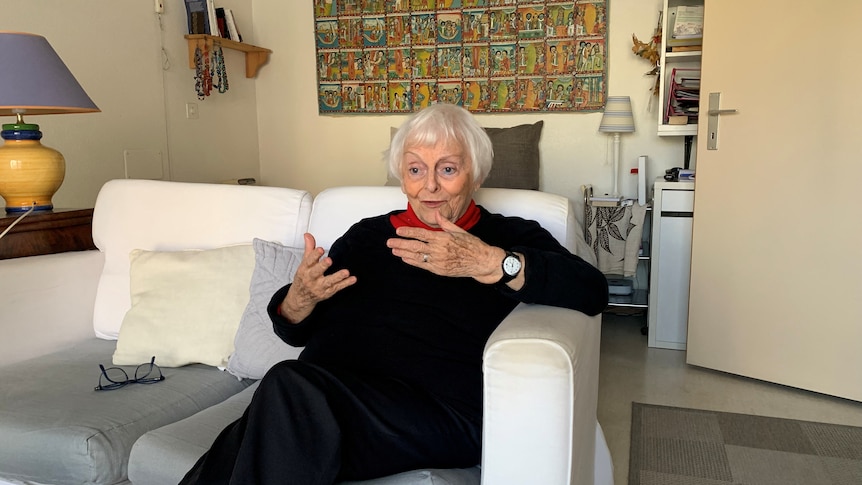 An elderly woman, sitting on a small white sofa in a living-room, gestures with her hands