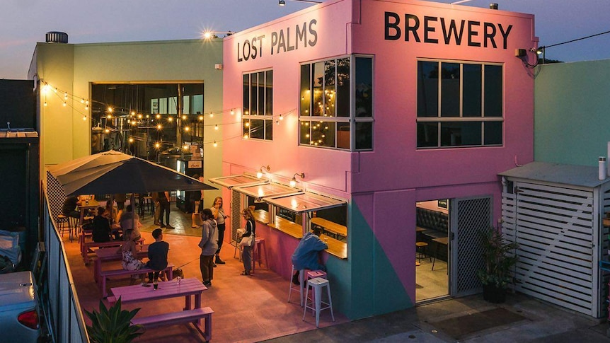 Early evening shot of a pink building with the writing that says Lost Palms Brewery. Seat with people are outside the building.