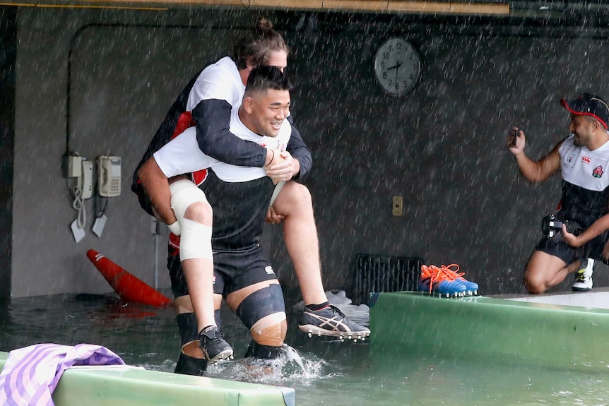 A big rugby player carries another rugby player on his back as they wade through knee-high water.