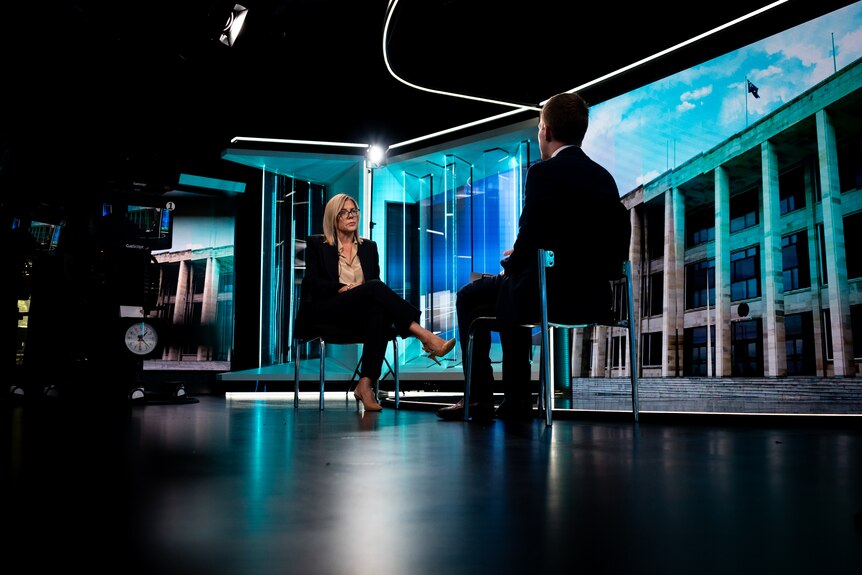 A wide shot of Libby Mettam in a dark pantsuit sitting in a television studio being interviewed by Keane Bourke.