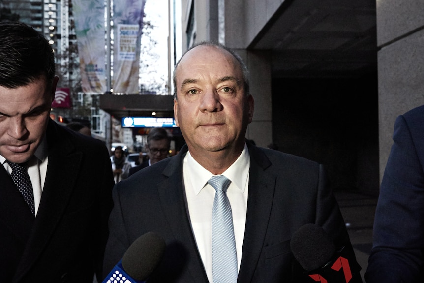 Daryl Maguire walks as two journalists hold microphones to him on either side