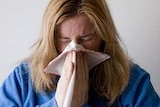 A women with long blonde hair and a blue jumper sneezes in to a tissue
