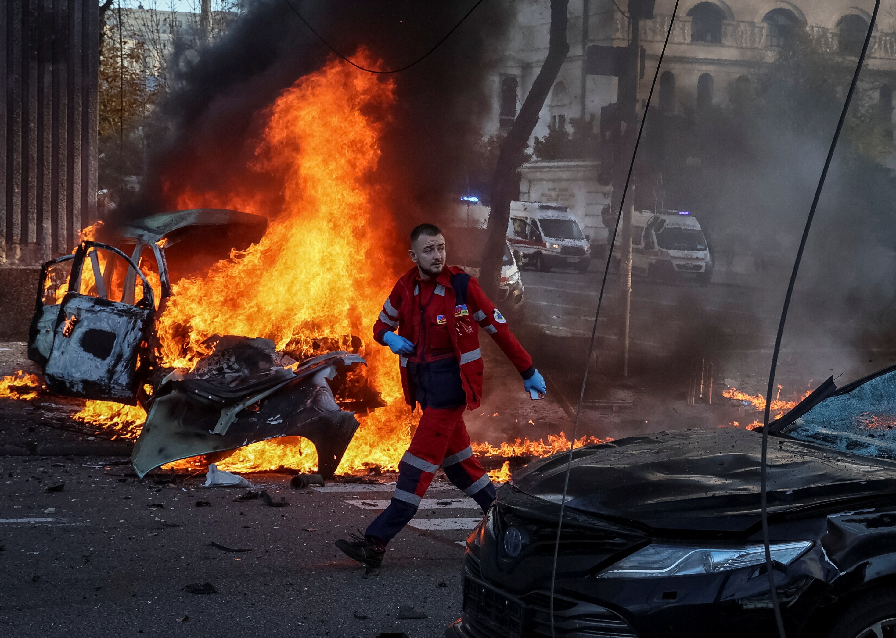 A medical worker surveys damage in Ukraine. A car is burning behind him. 