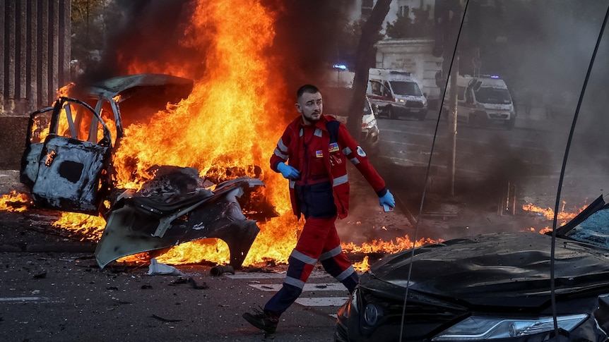 A medical worker surveys damage in Ukraine. A car is burning behind him. 