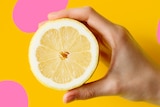 A white hand holds a lemon cut in half against a yellow background with light pink spots.