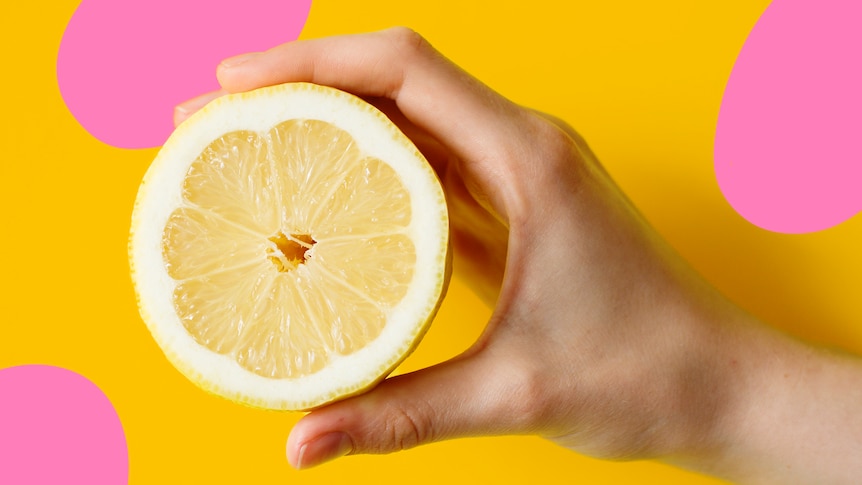 A white hand holds a lemon cut in half against a yellow background with light pink spots.