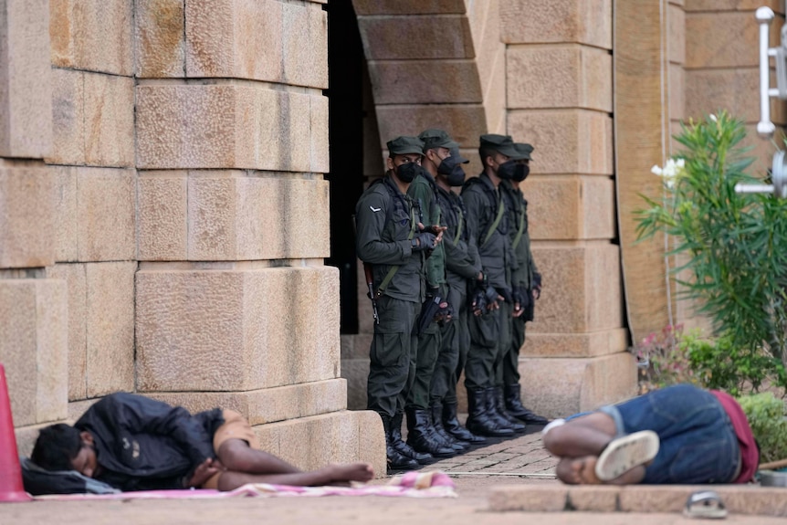 A group of soldiers guard a building as two people sleep on the ground nearby