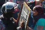 A police officer in riot gear holds his shield against a woman in a crowd