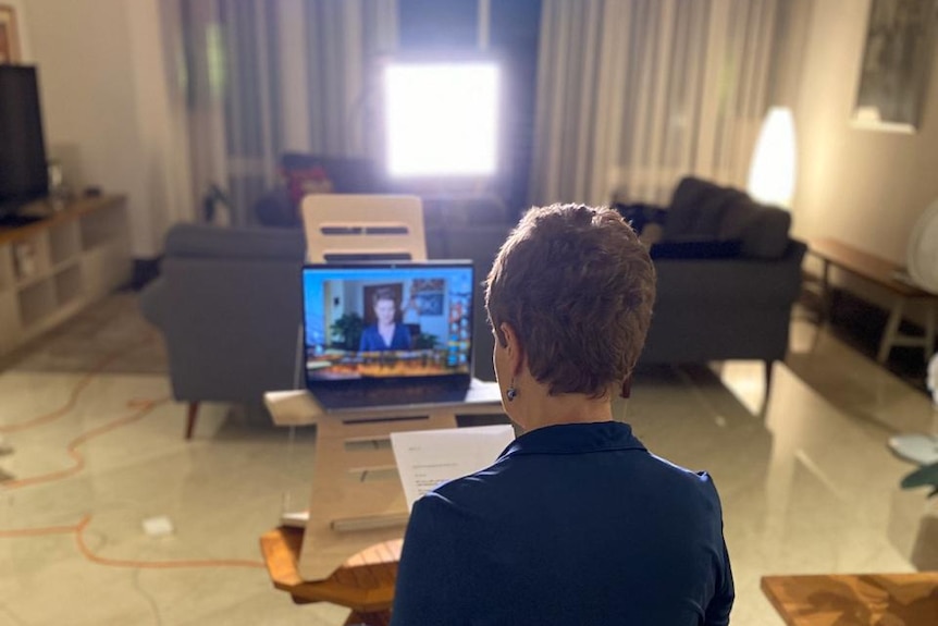 Barker in lounge room sitting on chair looking at laptop with her face on screen.