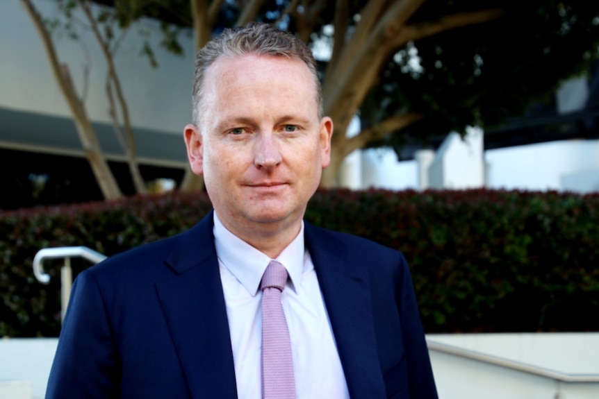 A mid shot of a man wearing a mauve tie, white shirt and navy blue blazer standing with a tree in the background.