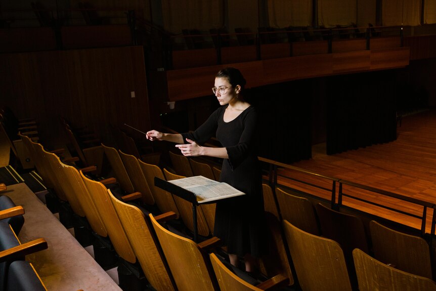 Wide shot of Nicky Gluch conducting with hands hovered over music sheet in concert hall.