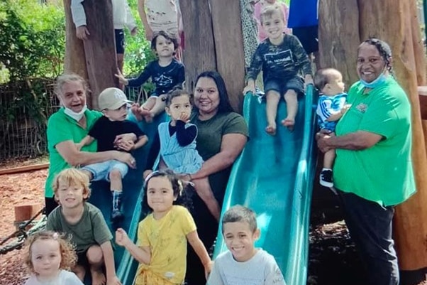 Photo of children sitting on and around play equipment with three adults standing with them, smiling.