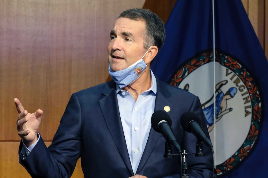 A man gestures as he speaks at a podium with a facemask pulled under his chin