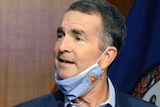 A man gestures as he speaks at a podium with a facemask pulled under his chin