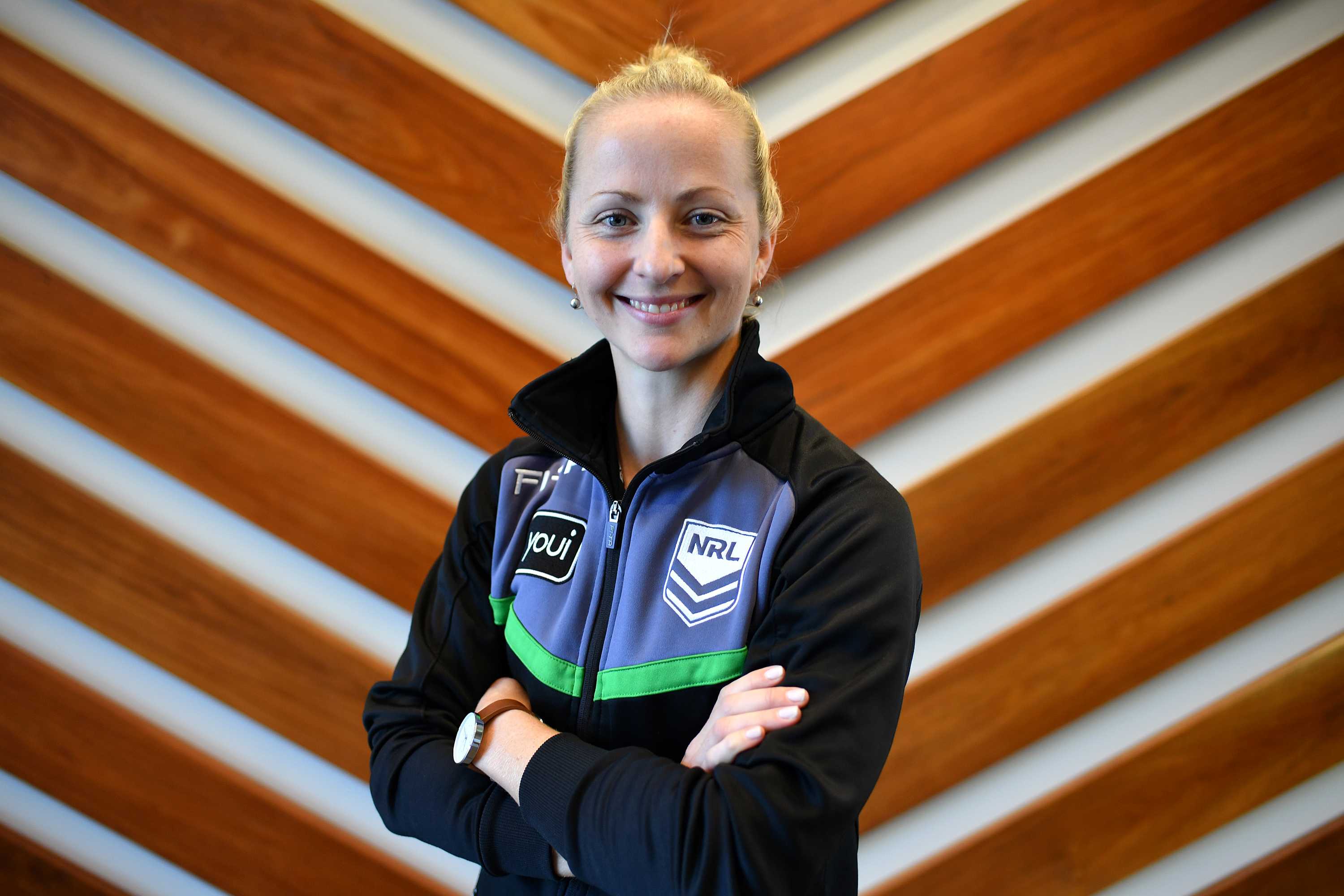 A woman with blonde hair pulled into a ponytail and wearing a referee jacket smiles with her arms crossed.