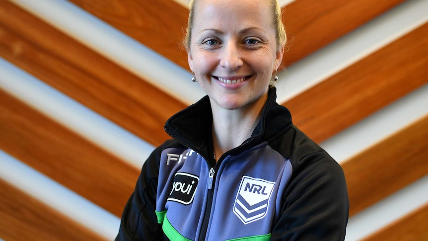 A woman with blonde hair pulled into a ponytail and wearing a referee jacket smiles with her arms crossed.