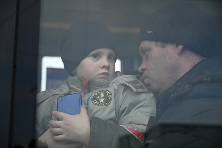 A young boy holding a phone and sitting on a man's lap inside a bus looks out of the bus window.