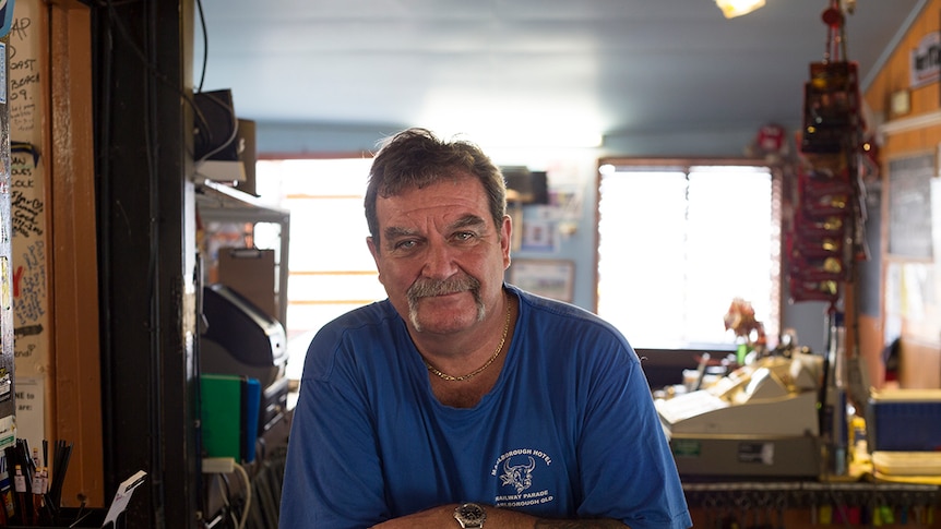 A man stares directly at the camera, leaning on a bar.