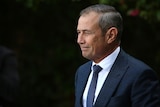 A side view of Roger Cook who is smiling and wearing a suit
