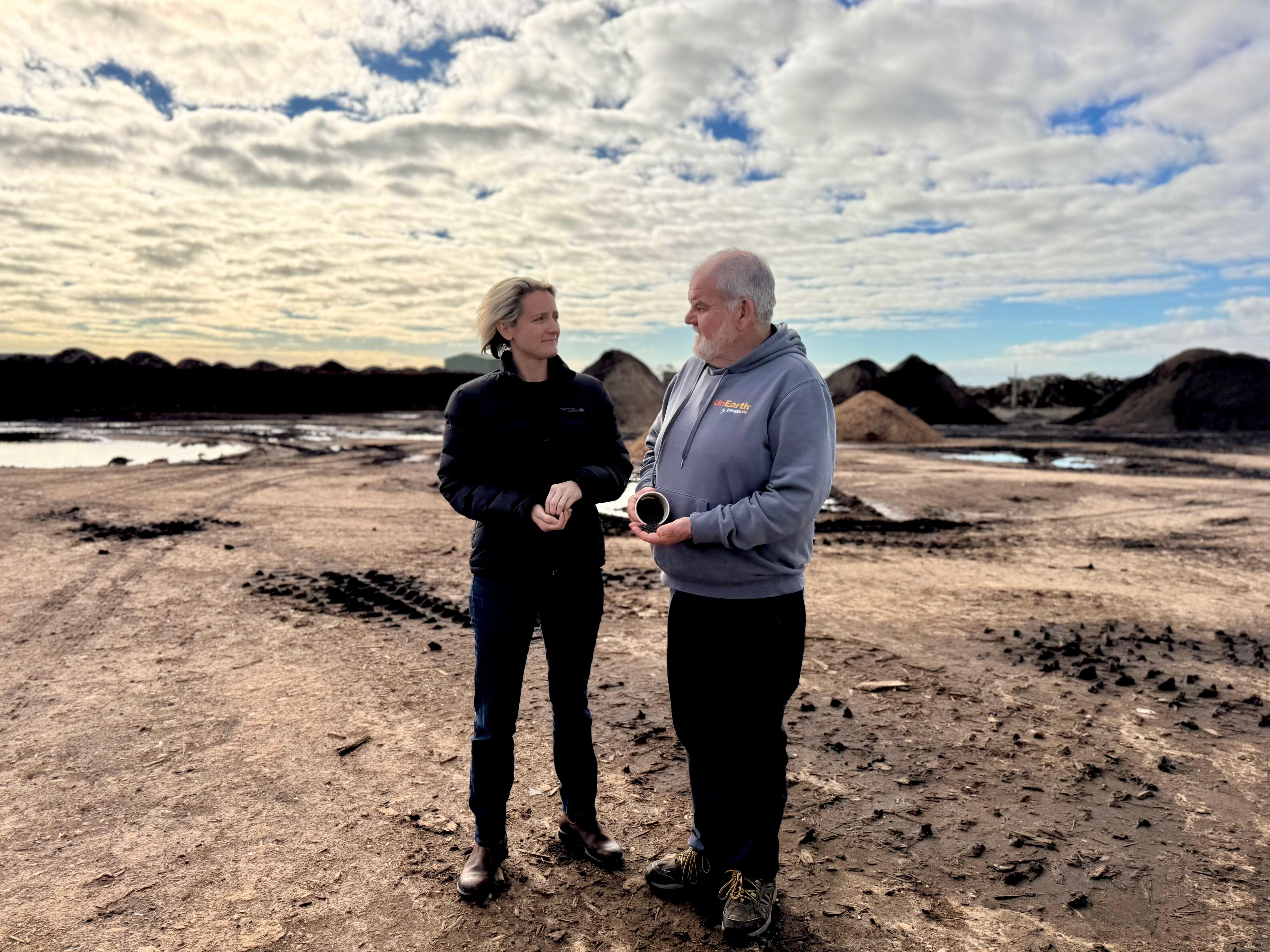 Two people talk to each other while standing on dirt and holding soil samples.
