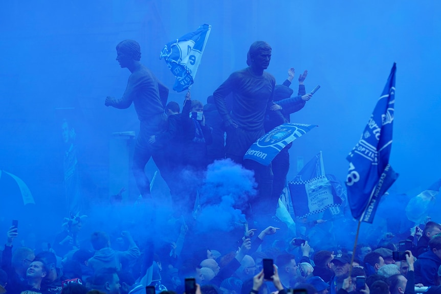 A statue outside Everton's stadium is shrouded in blue smoke as fans wave flags and film on their cell phones.