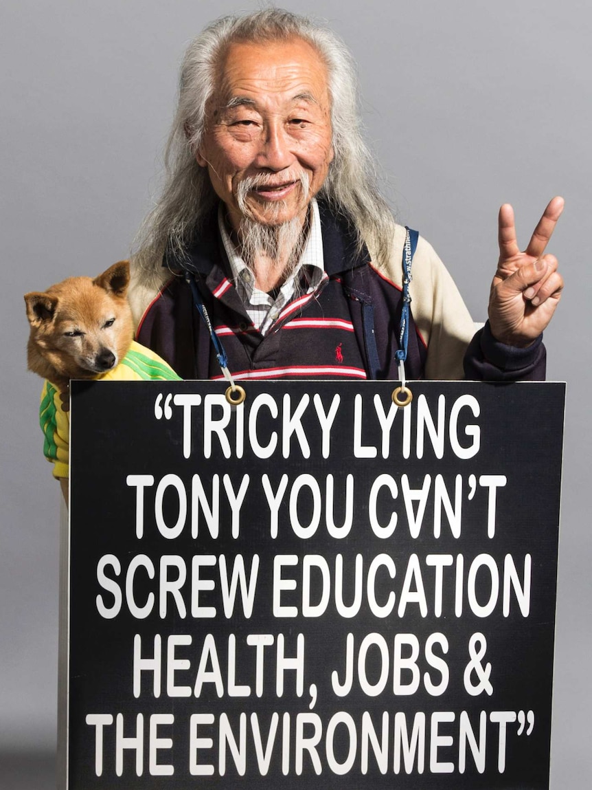 Old man with dog in hand does peace sign with his fingers, while holding a placard criticising Tony Abbott.