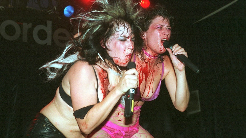 two women in underwear scream into microphones. both their chests are covered in blood