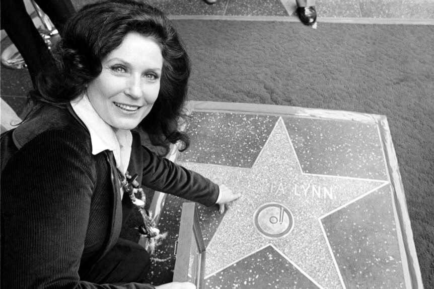 black and white, woman with dark hair smiles at the camera, crouching, and points to walk of fame star