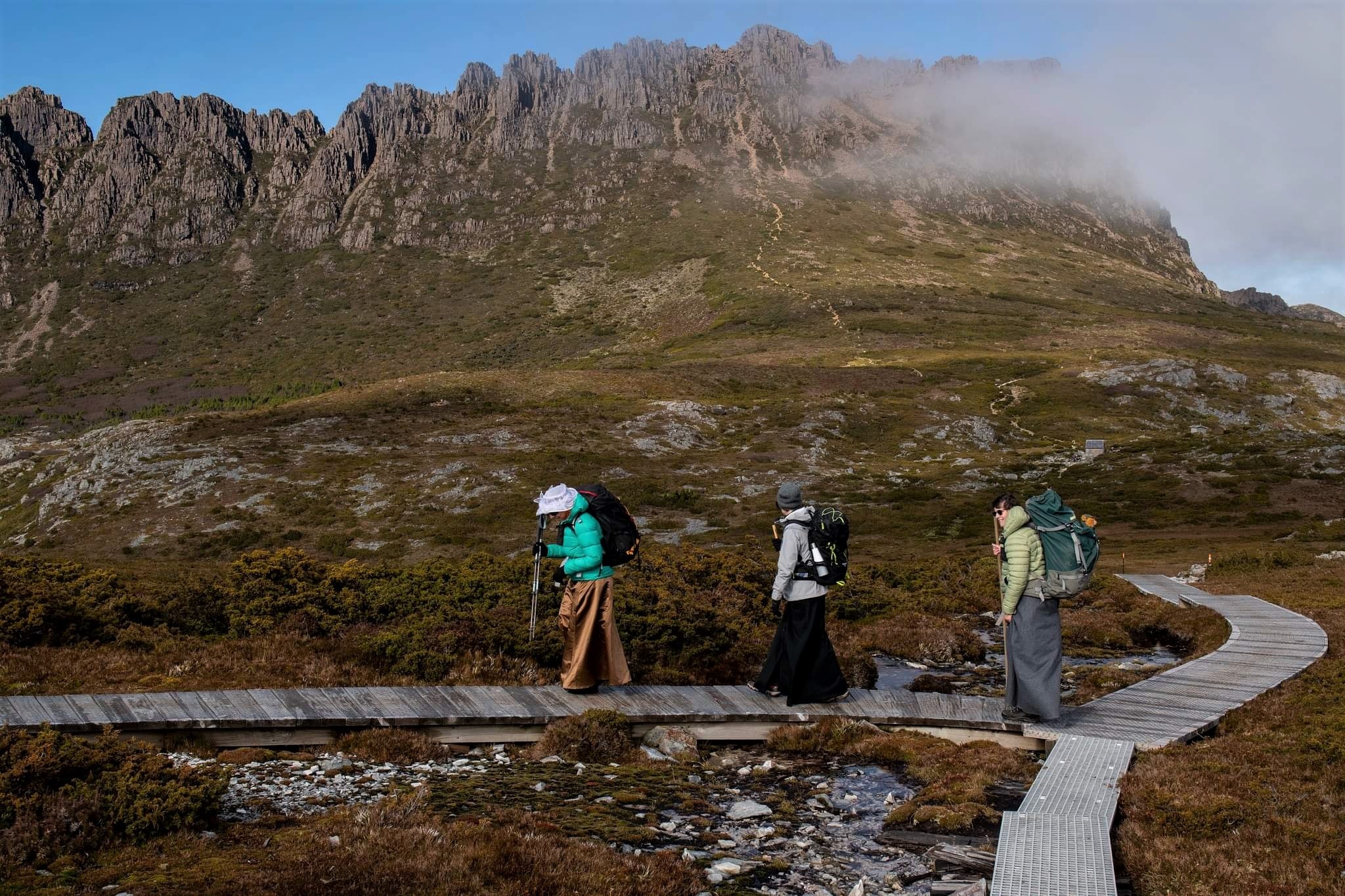 Three women wearing long skirts and puffer jackets, walking with Cradle Mountain's peak behind them.