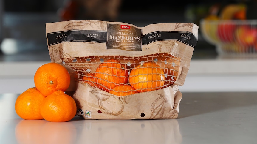 Mandarins loose on a kitchen counter next to mandarins packed in a Coles-branded brown recyclable paper bag with a net window. 