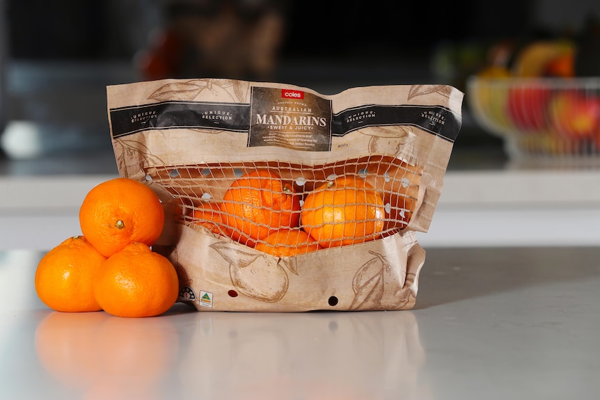 Mandarins loose on a kitchen counter next to mandarins packed in a Coles-branded brown recyclable paper bag with a net window. 