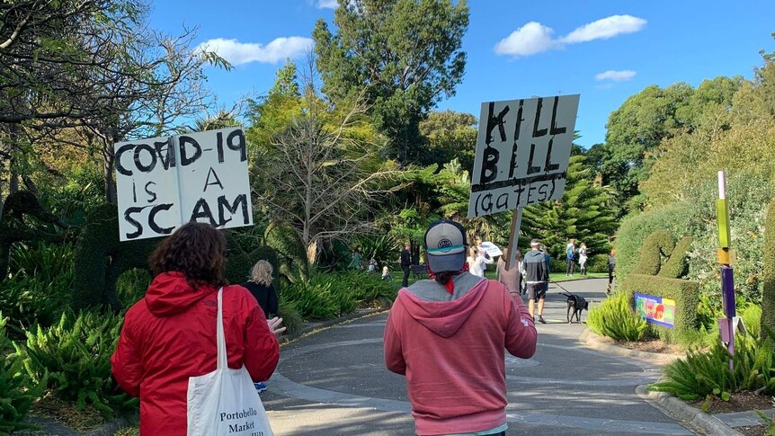 A shot from behind of two protestors holding signs. One says: "COVID-19 is a scam", the other says "KILL BILL (Gates)"