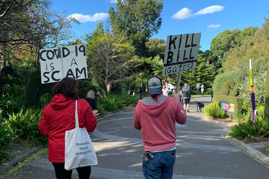 A shot from behind of two protestors holding signs. One says: "COVID-19 is a scam", the other says "KILL BILL (Gates)"