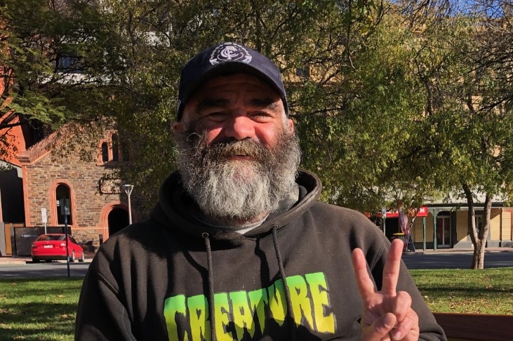 A bearded man wearing a baseball cap makes a peace sign with a sign saying 'smile it's free'.