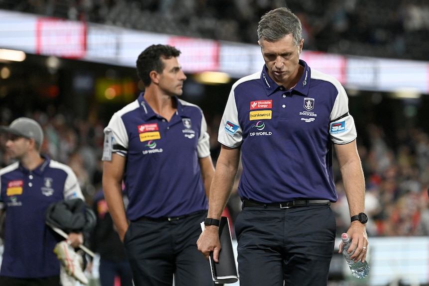 A man in a purple polo shirt with his head bowed walking with another man behind him. 