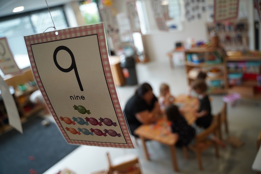 Unidentified teacher and children sit at a desk in a day care centre.