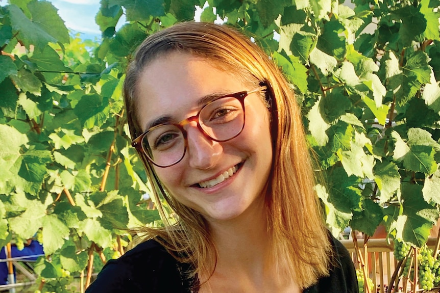 A young woman in glasses smiles at the camera in front of grapevine leaves