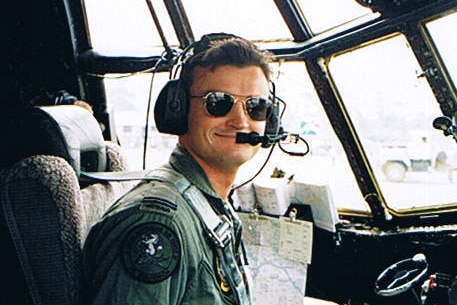 A young man sits in the cockpit of an Air Force plane with a headset and sunglasses on smiling for a photo.