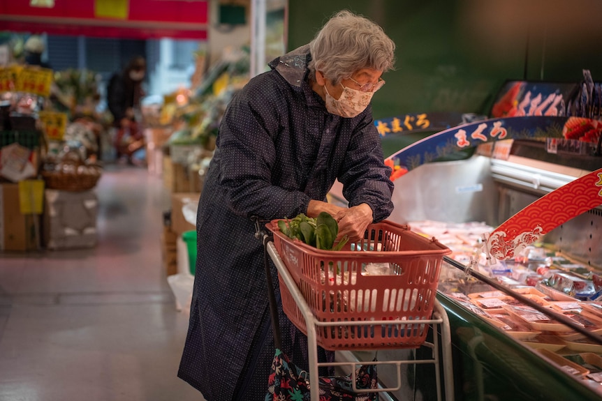 An elderly woman leans over a shopping trolley with a basket on top, next to a deli counter