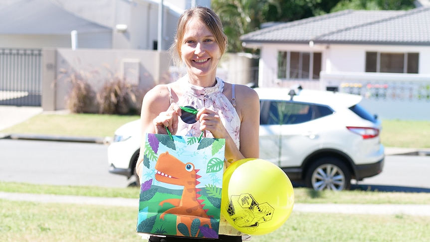 woman with light brown hair in a front yard holds a dinosaur party bag and yellow balloon with a white car in the background