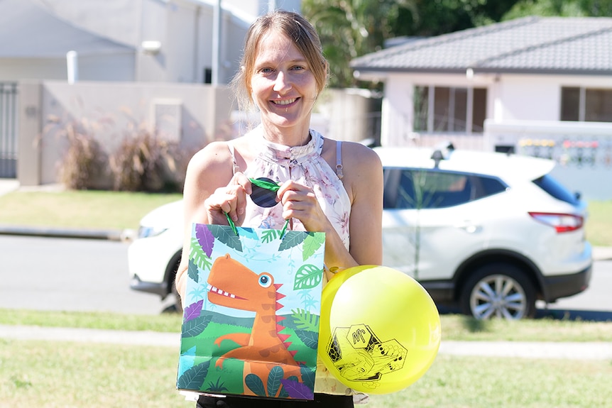 woman with light brown hair in a front yard holds a dinosaur party bag and yellow balloon with a white car in the background